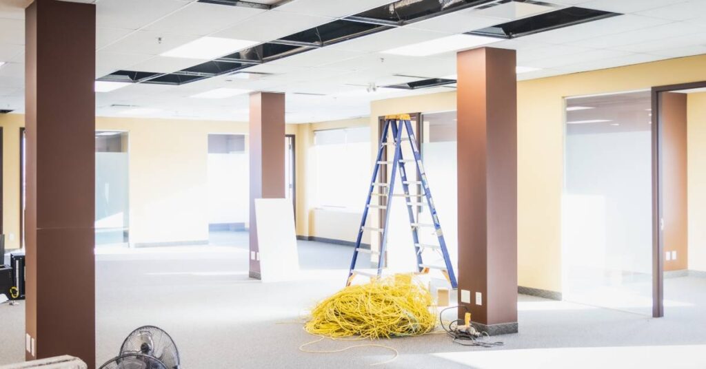 A large office space is experiencing renovations. There is a ladder in the background and several ceiling tiles removed.