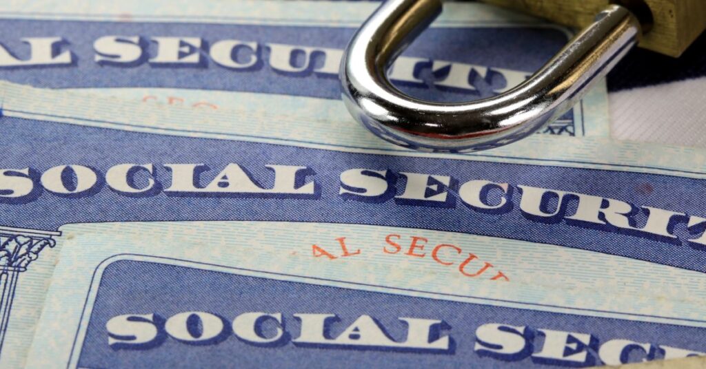 Three Social Security cards are strewn across a table with a gold and silver padlock laying across them.
