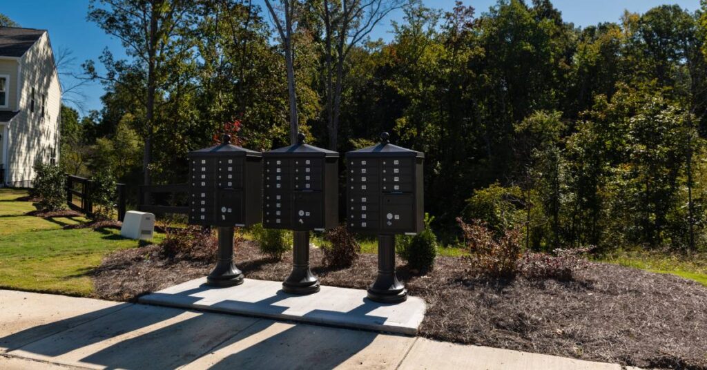 Three black cluster mailboxes rest along a street on a sunny day with grass and a grove of trees behind them.