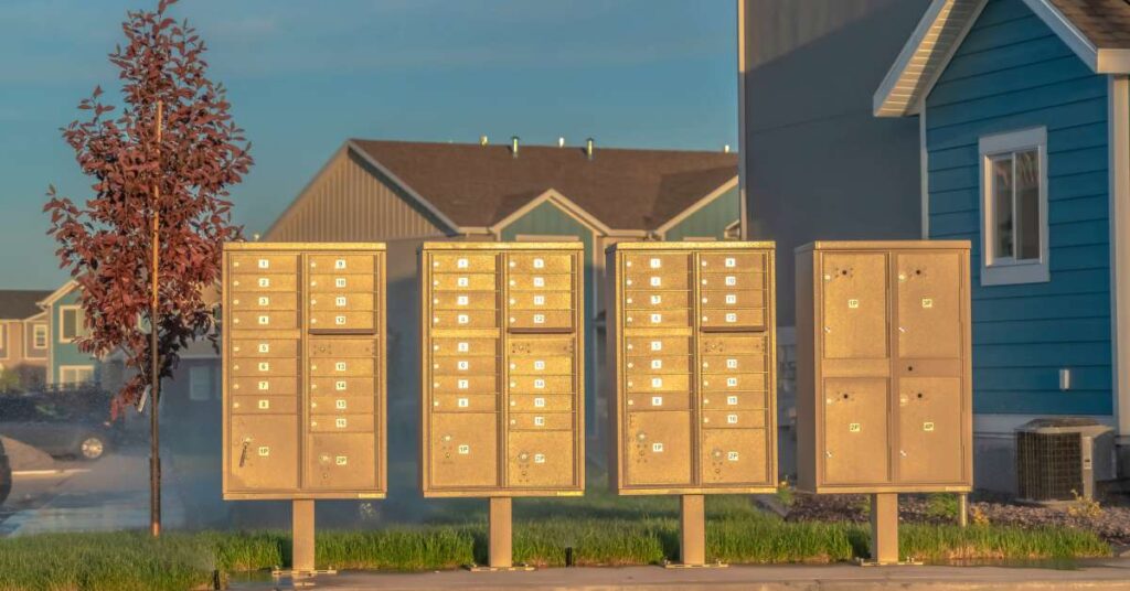 Four sets of golden brown cluster mailboxes surrounded by houses sit on a curb while the sun sets in the distance.