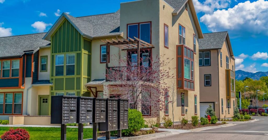 A row of gray cluster mailboxes stand in front of a modern-looking home on a bright day with clear blue skies. .