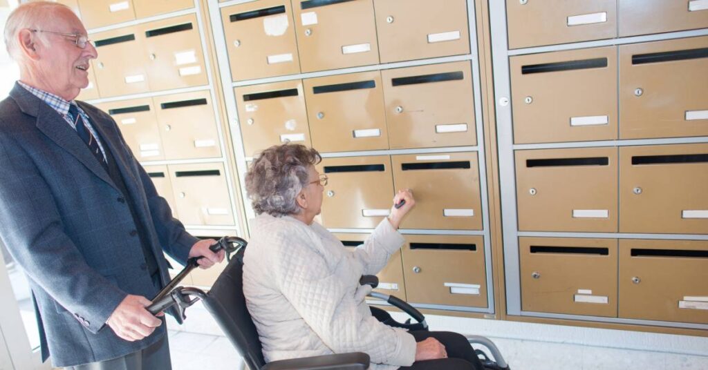 A man stands behind a woman as she sits in a wheelchair and locks or unlocks an individual mailbox compartment.