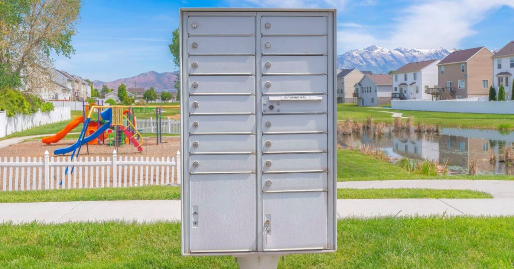 In the foreground is a cluster mailbox unit. In the background is a playground, a lake, and a few houses.