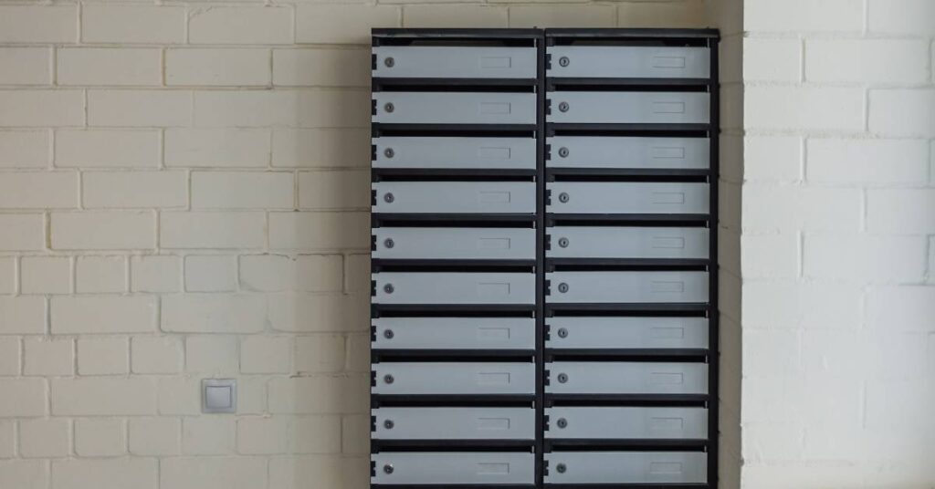A black and gray mailbox with twenty lockable compartments is attached to a white brick wall on a property.
