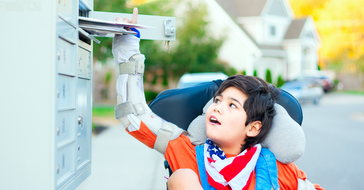A boy sitting in a wheelchair, with his arms in casts, looks up as he reaches into a community mailbox.