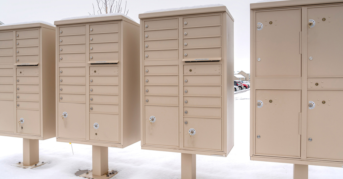 Four beige cluster mailbox units sit outside on snow-covered ground between multicolored residential buildings.