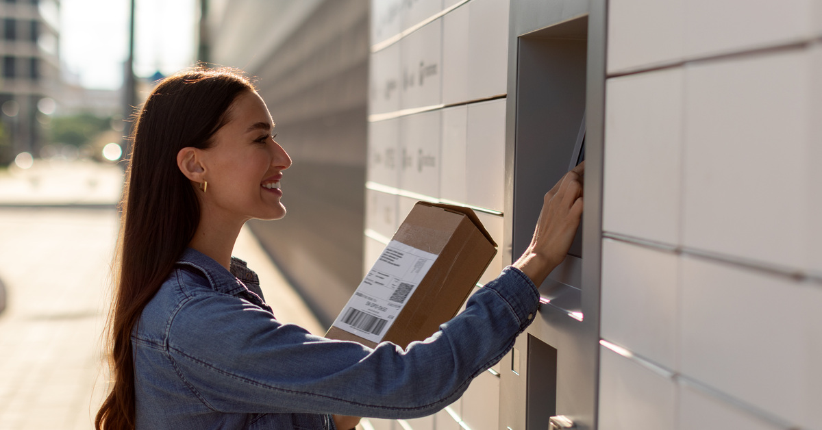 A woman smiles as she holds a cardboard box and uses the touchscreen display on an outdoor mailbox system.