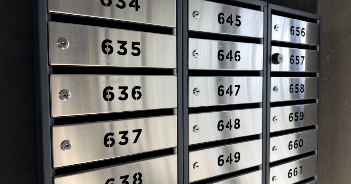 Row of modern stainless steel mailboxes with bold black numbers and built in locks mounted on dark wall.