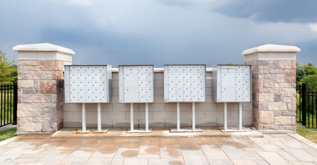 Cluster mailbox units installed against a brick wall with black gates on either side and trees in the background.