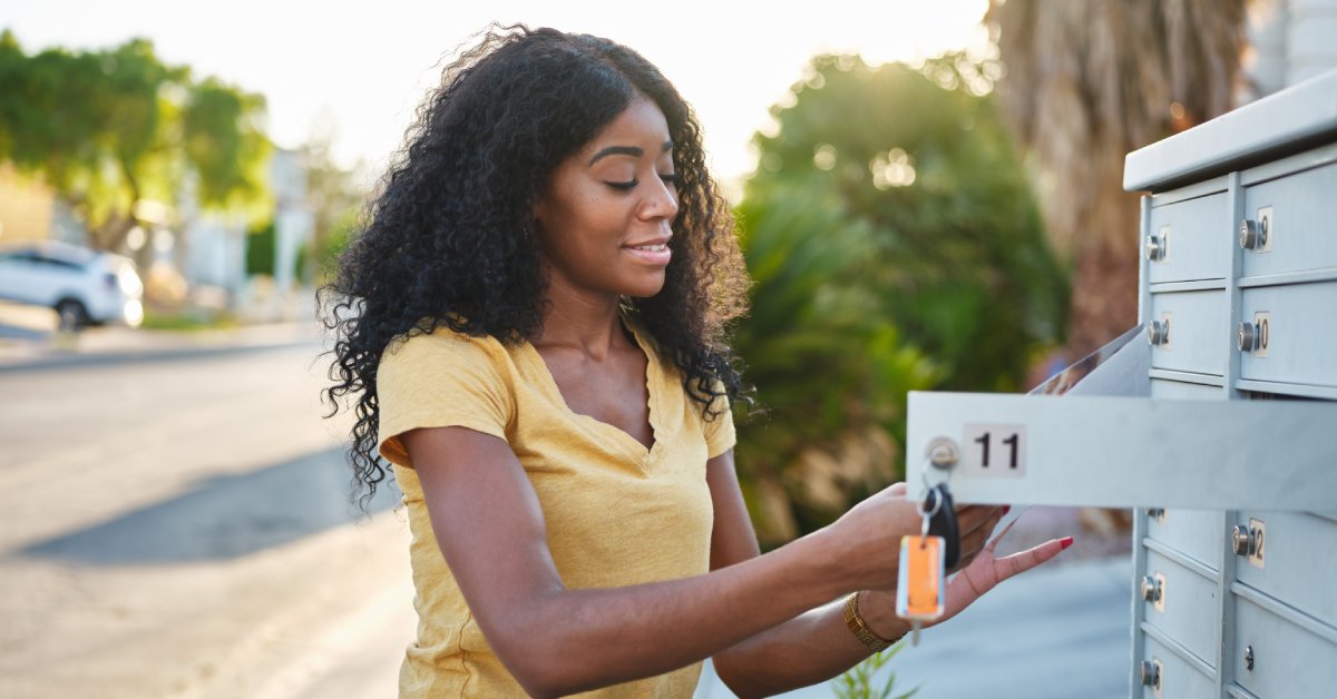 A woman in a yellow shirt stands in front of a cluster mailbox, where a key is inside the lock of an opened individual slot.