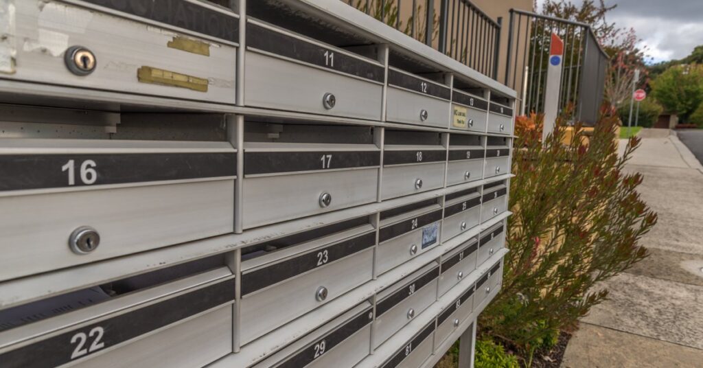A community mailbox unit with numbered slots, installed outside along a sidewalk with plants, near a gated building.