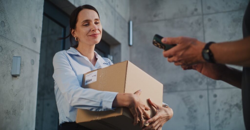 A woman holding a brown cardboard box and facing another person who holds a mobile device in their hands.