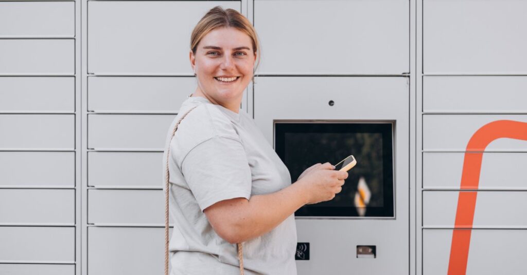 A woman smiling as she holds a mobile phone while standing in front of an automated mailbox and parcel machine.