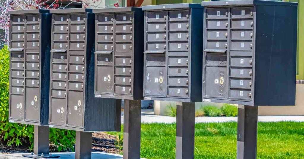 A row of neutral-colored cluster mailboxes with numbered individual units installed on a sidewalk near grass.