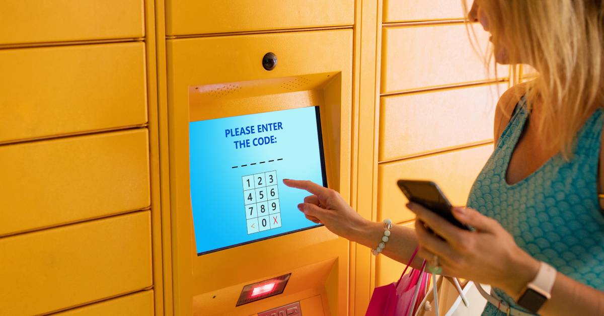 A woman holding a cell phone stands in front of a parcel locker with a digital screen that prompts her to enter a code.