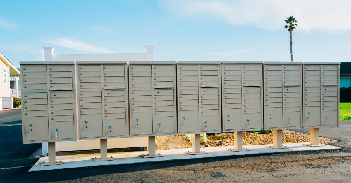 A row of lightly colored cluster mailbox units made of metal installed on a sidewalk in a neighborhood.