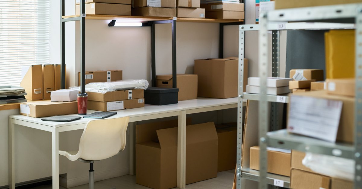 A room with a desk and a shelving unit in the foreground. On the desk there are Several boxes. There is a window by the desk.