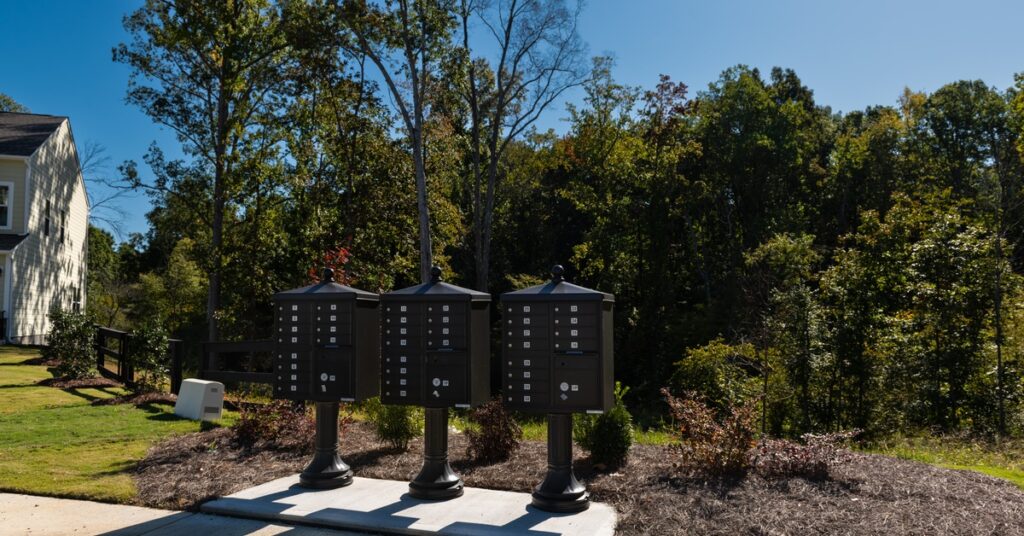 Three cluster mail boxes erected along the sidewalk. There are trees in the background behind them.