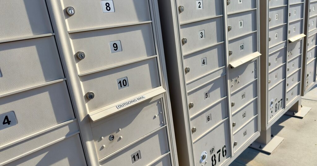 Mailboxes are on a concrete surface. The mailboxes themselves are steely gray. They are outside and sunlight is on them.