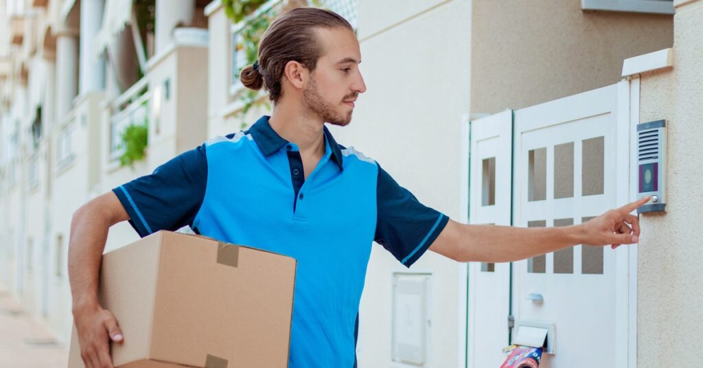 A delivery man in a blue shirt is holding a box under one arm. He is pressing a doorbell with his other hand.