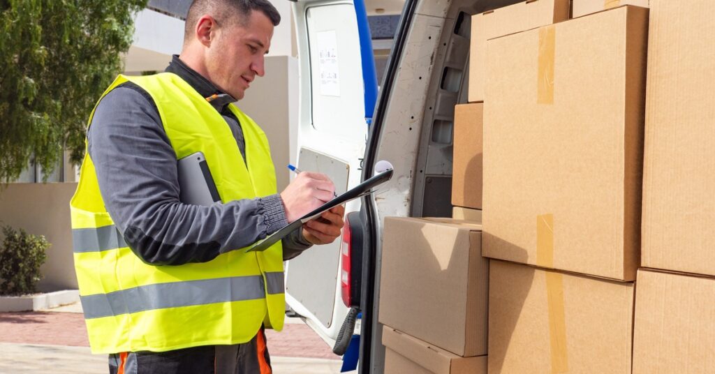 A man is holding a clipboard in one hand and a pen in the other. He’s standing in front of a van with boxes in it.