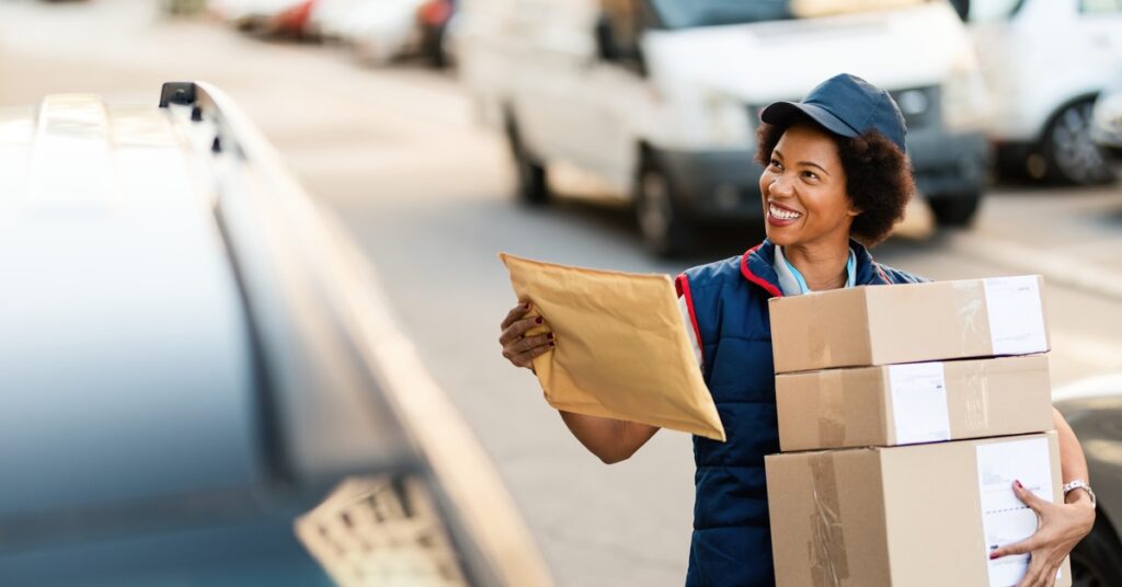 A woman in a mail carrier’s outfit is holding a stack of cardboard boxes in her arms. She is smiling.