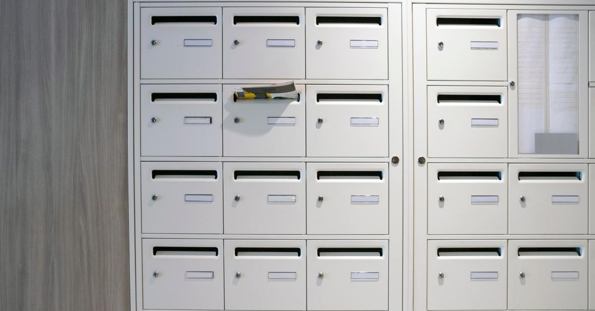 There are mailboxes attached to a wall with a directory to the right of the frame. Some mail is hanging out of one of the boxes.