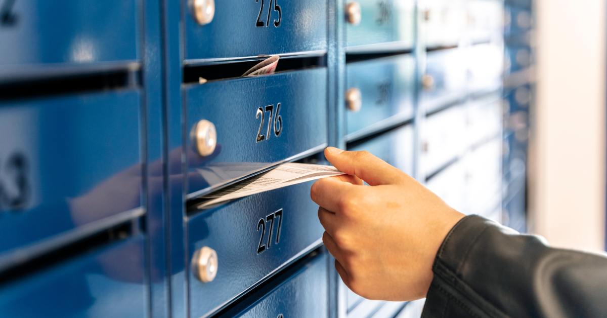 A hand is grabbing a letter that is sticking out of a blue mailbox. The wall is filled with blue mailboxes.