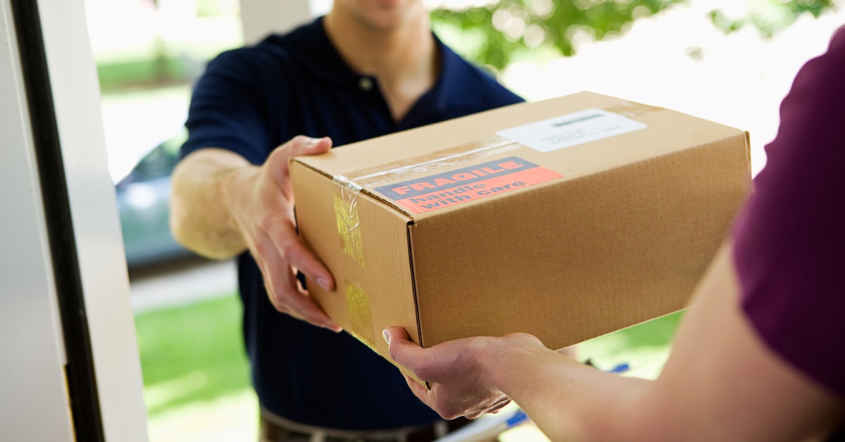 A man is handing a box to a person. The man delivering the box is wearing a blue shirt tucked into his pants.